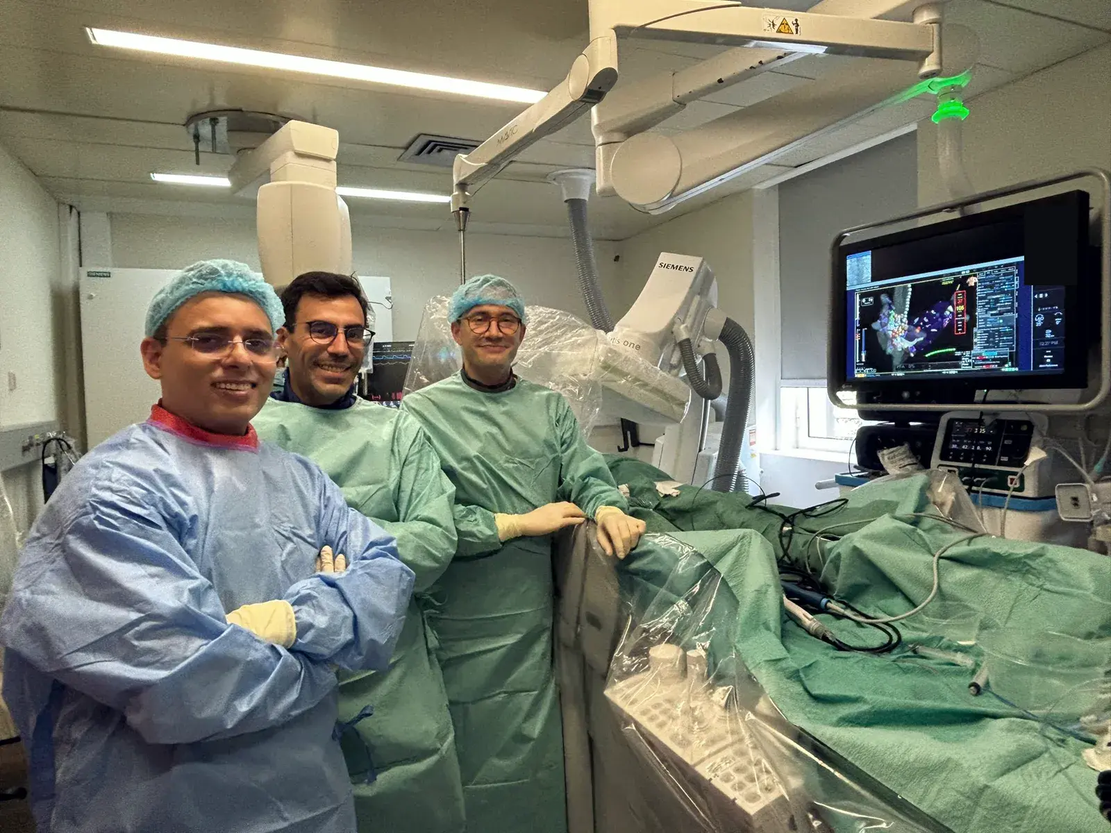 Three physicians in surgical attire inside an electrophysiology laboratory; a monitor displays a three-dimensional electroanatomic map of the heart.