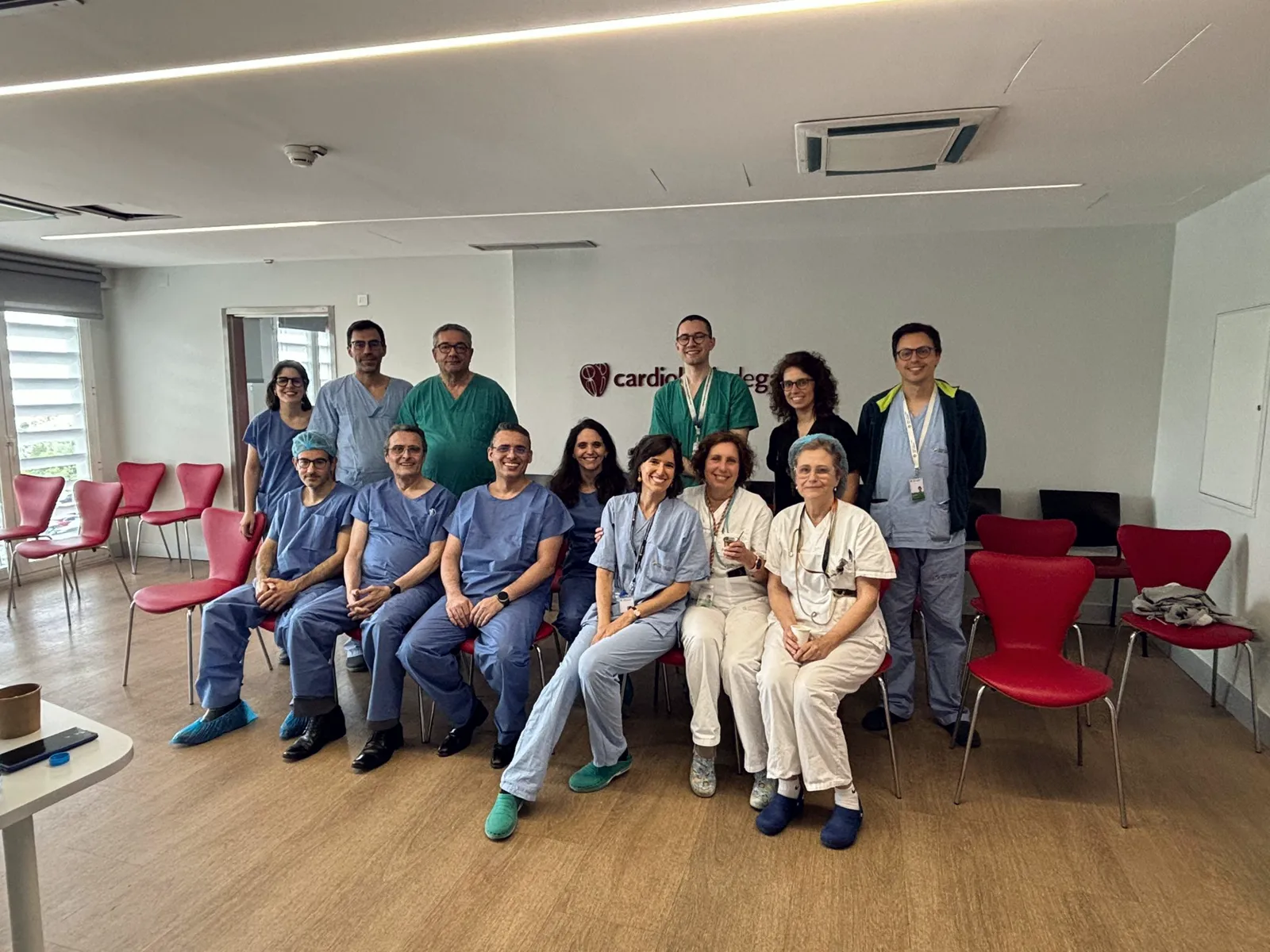 Group photograph of physicians in scrubs and white coats, seated and standing in a hospital meeting room during a proctoring course.