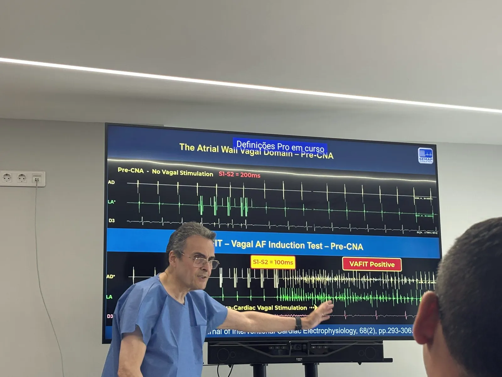 Physician in blue scrubs pointing at a large screen showing ECG and intracardiac tracings labeled 'VAFIT — Vagal AF Induction Test' and 'VAFIT Positive.'
